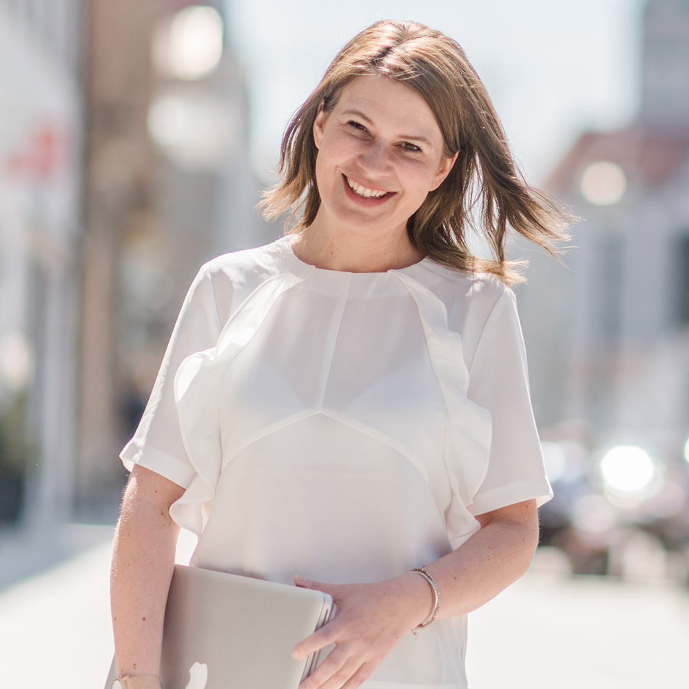 Bernadette lächelt mit ihrem Laptop in der Hand in die Kamera